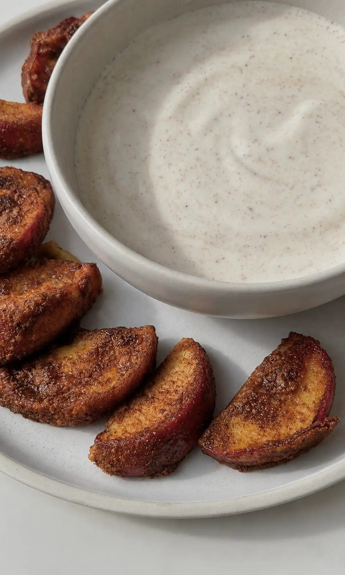 Air fryer apple fries coated in crisp cinnamon coconut sugar on a speckled ceramic plate next to a bowl of creamy cinnamon yogurt dip