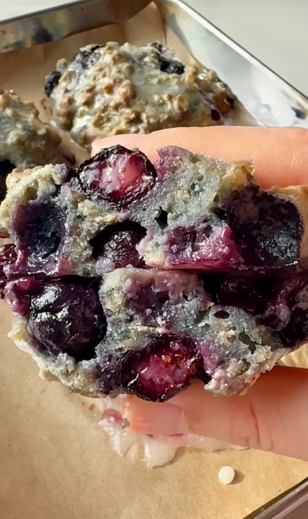 A hand holding a blueberry fritter broken in half showing the jammy blueberry filling inside with more fritters and a baking tray in the background