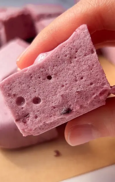 A hand holding a piece of pink cherry marshmallow showing its soft, spongy texture, with more pieces on a wooden board in the background