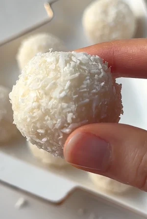 A hand holding a single healthy coconut truffle ball coated in shredded coconut