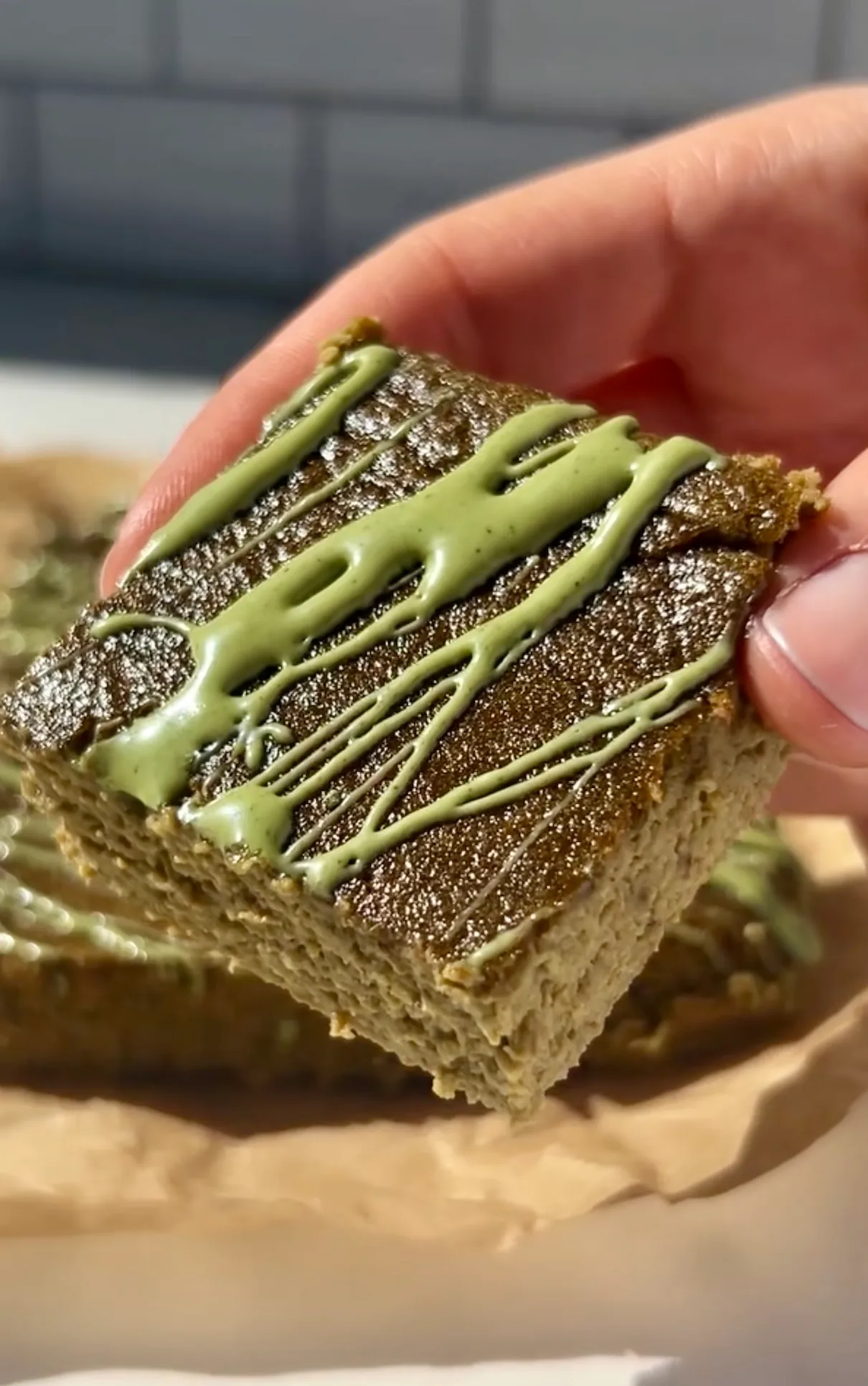A hand holding a square slice of easy healthy matcha cake close up showing the golden-green sponge with bright green matcha icing drizzled on top against a white tile background