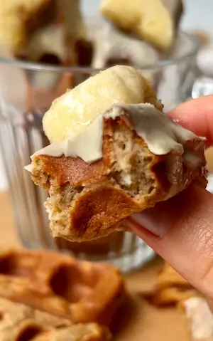 A hand holding a gluten free banana bread waffle stick with white chocolate dripping down and a banana slice on top