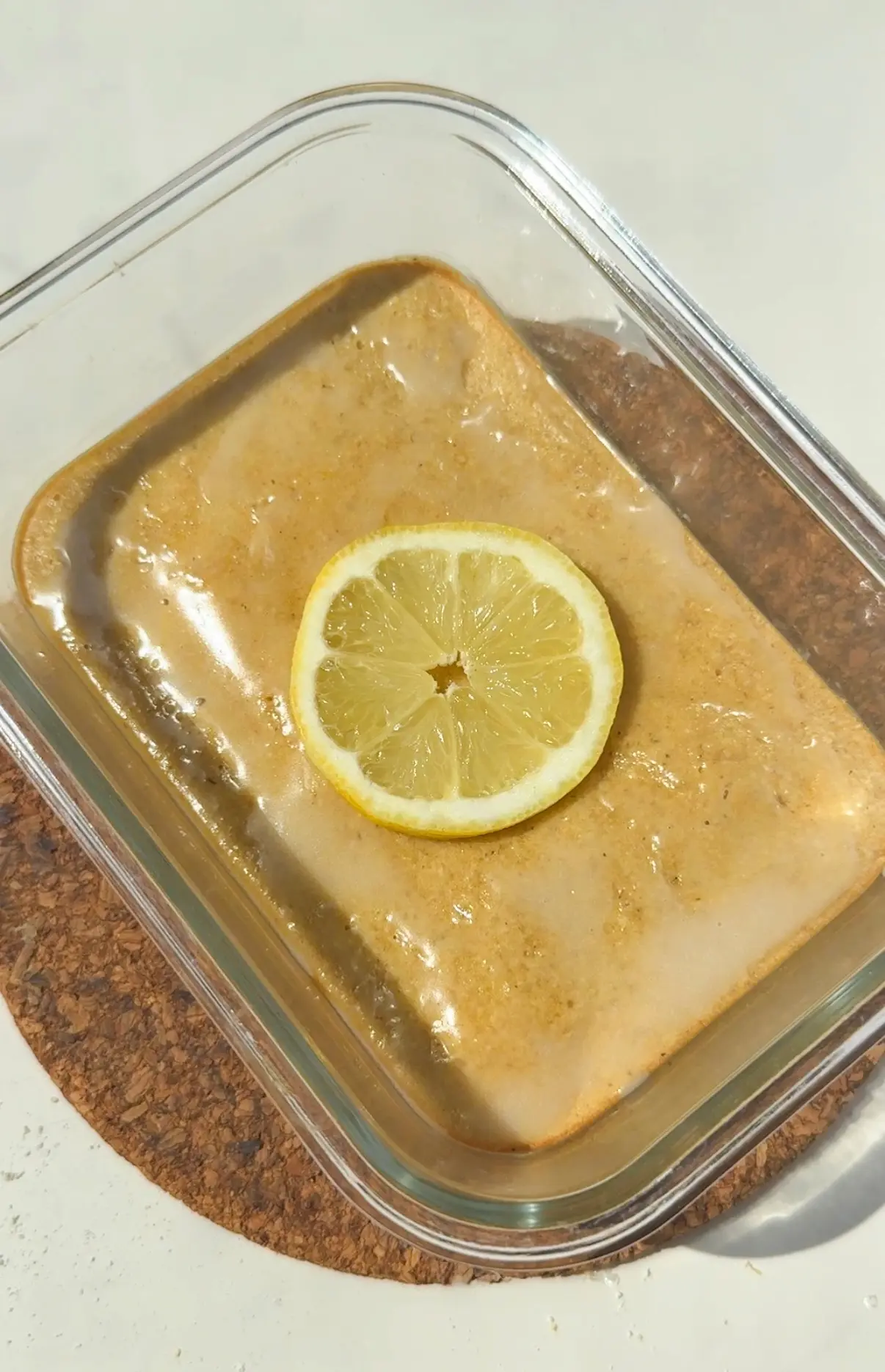 A golden lemon drizzle pancake bowl topped with a white drizzle in an oven-safe bowl, with lemon zest and a slice of lemon on the side