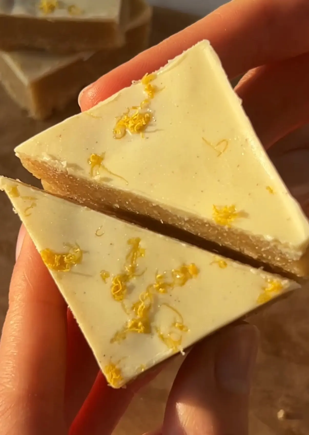 A hand holding two no-bake lemon bars cut into triangles showing the golden almond crust and creamy white chocolate lemon layer topped with lemon zest
