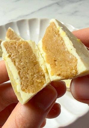 A hand holding a no bake lemon cup broken in half showing the dense lemon almond flour filling inside a white chocolate shell