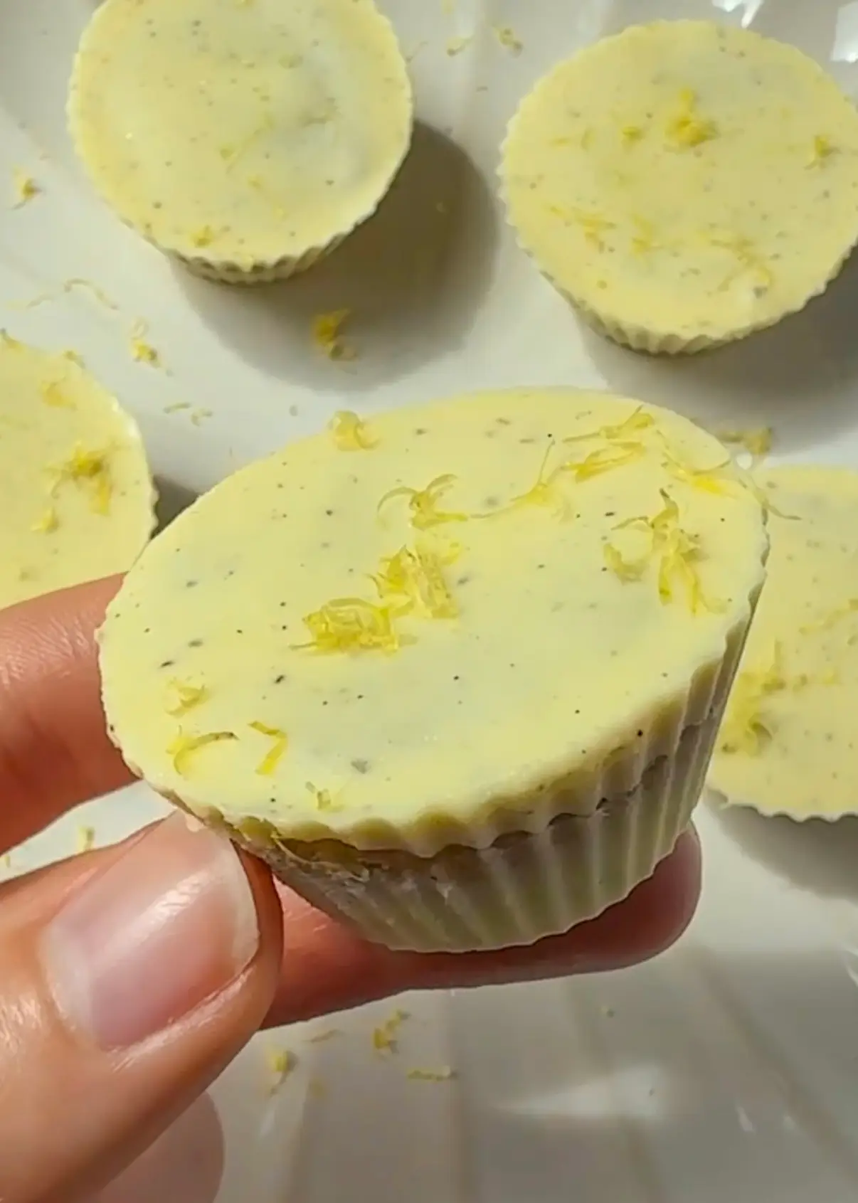 A hand holding a single no bake lemon cup topped with lemon zest with more cups on a white plate in the background