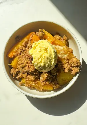 Overhead view of healthy peach crumble in a bowl with golden oat topping, peach slices, and a scoop of vanilla ice cream