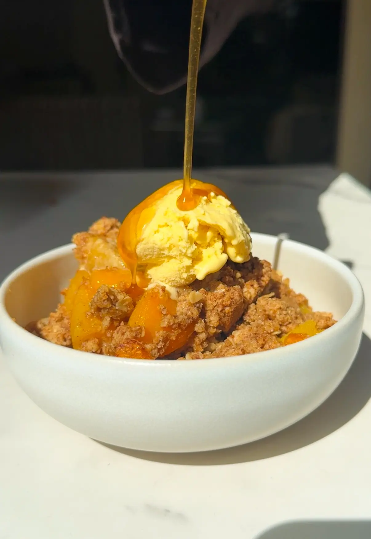 Healthy peach crumble in a bowl topped with vanilla ice cream and honey being drizzled over the top