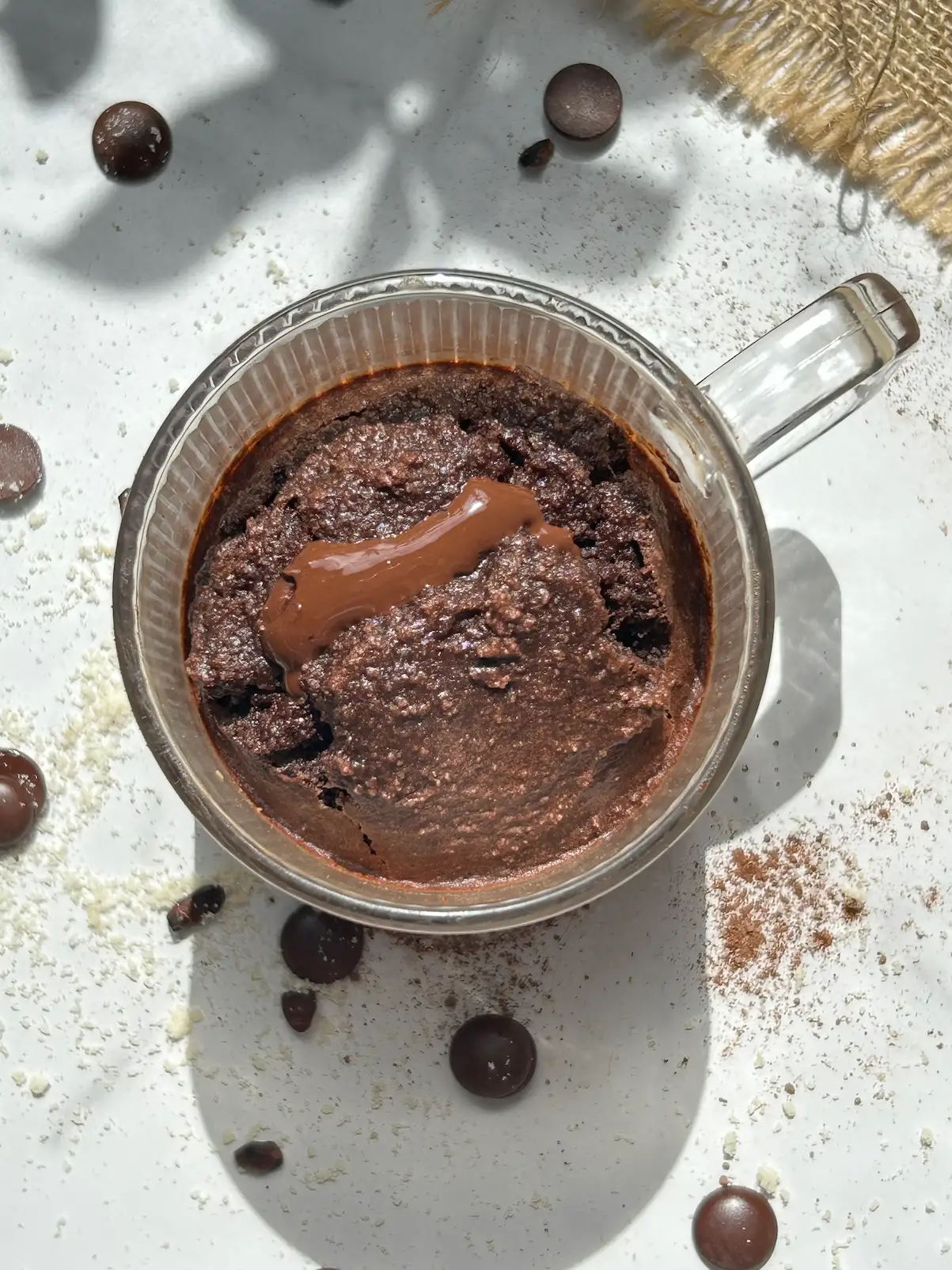 Overhead view of a triple chocolate mug cake in a glass mug with a glossy molten chocolate centre, surrounded by scattered chocolate chips, cocoa powder and a piece of burlap cloth